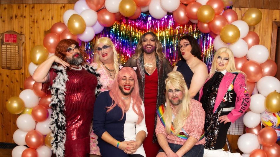 Seven grag queens posing in front of a rainbow ballon arch.