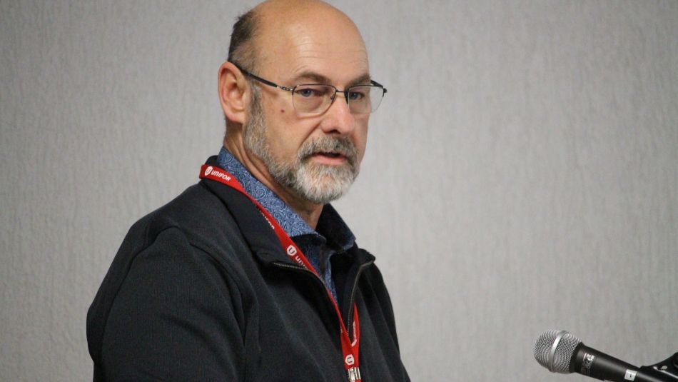 A man wearing a lanyard and dark top and glasses, speaks into a microphone.