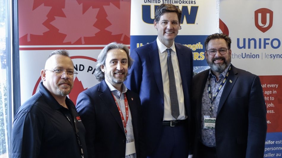 "Geoff Dawes, Gavin McGarrigle, le premier ministre David Eby et Scott Lunny posent pour une photo devant les bannières des syndicats.