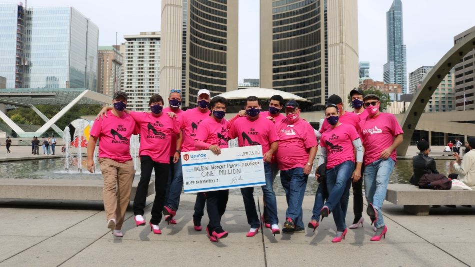 Unifor team at Hope In High Heels walk at Nathan Phillips Square in Toronto. Men are wearing pink high heels and holding a large cheque for 1 million dollars made out to Halton Women's Place