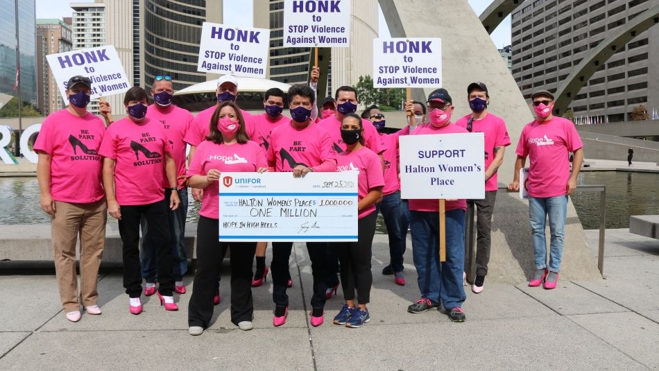 Unifor Hope In High Heels team with men wearing pink high heel shoes stand present large cheque to representatives from Halton Women's Place