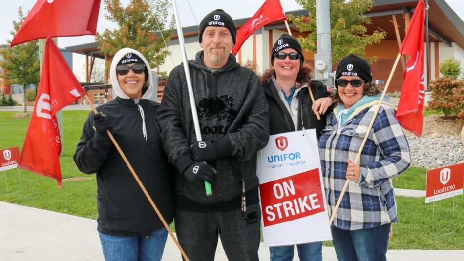 Four people posing for a photo outside with Unifor flags and an On Strike placard