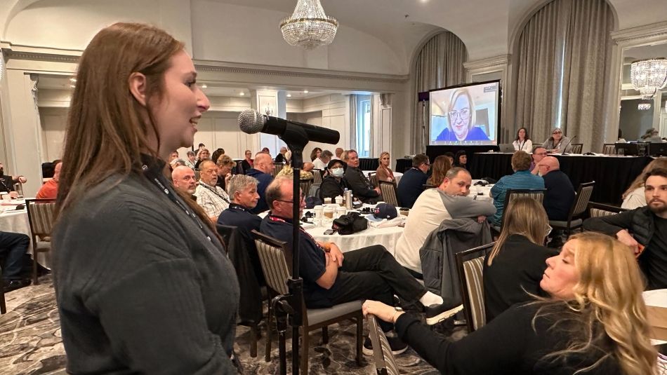 A woman at the podium speaks into a mic while a screen shows a woman on a Zoom call to a room full of people.