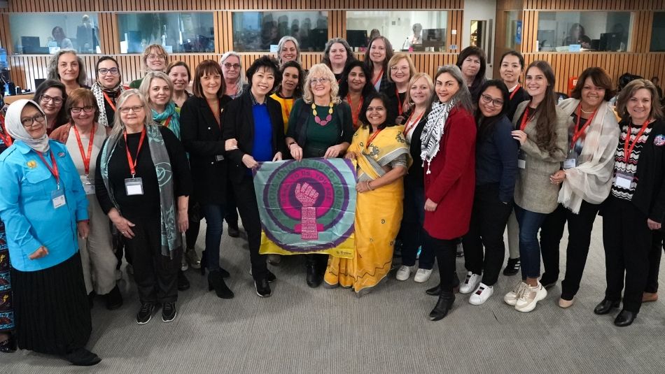 A diverse group of individuals standing together in an indoor setting, some wearing lanyards and badges, holding a circular banner with symbols and text advocating for rights and solidarity. The interior has wood paneling and glass windows in the background.