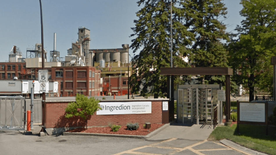 The entranceway of a factory with turnstiles and signage that reads, “Ingredion” with red brick building in the left background and industrial silos.