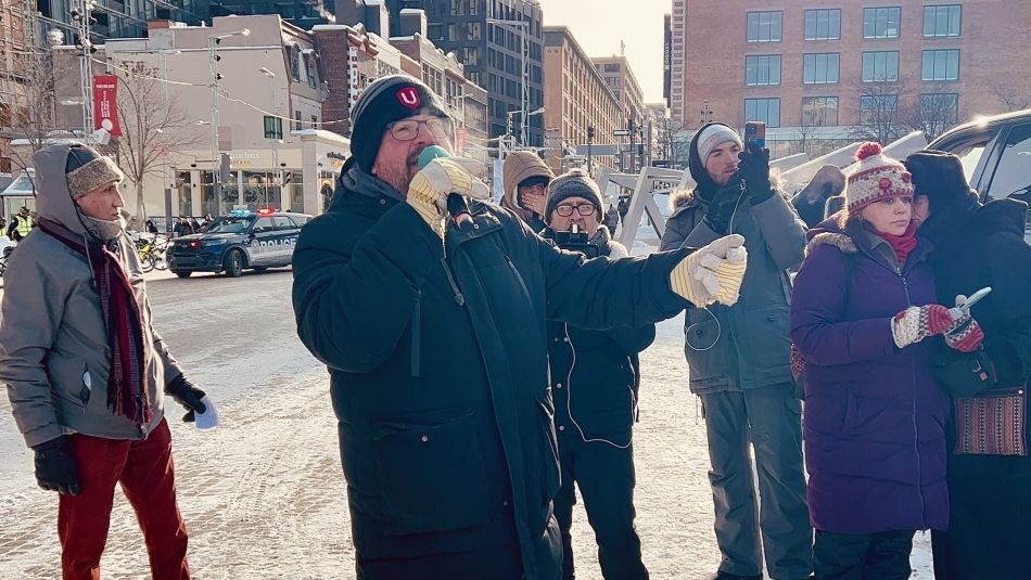 A man wearing a Unifor toque holds a mic and speaks to a crowd gathered around him on the street. Some people are taking photos using their phones.