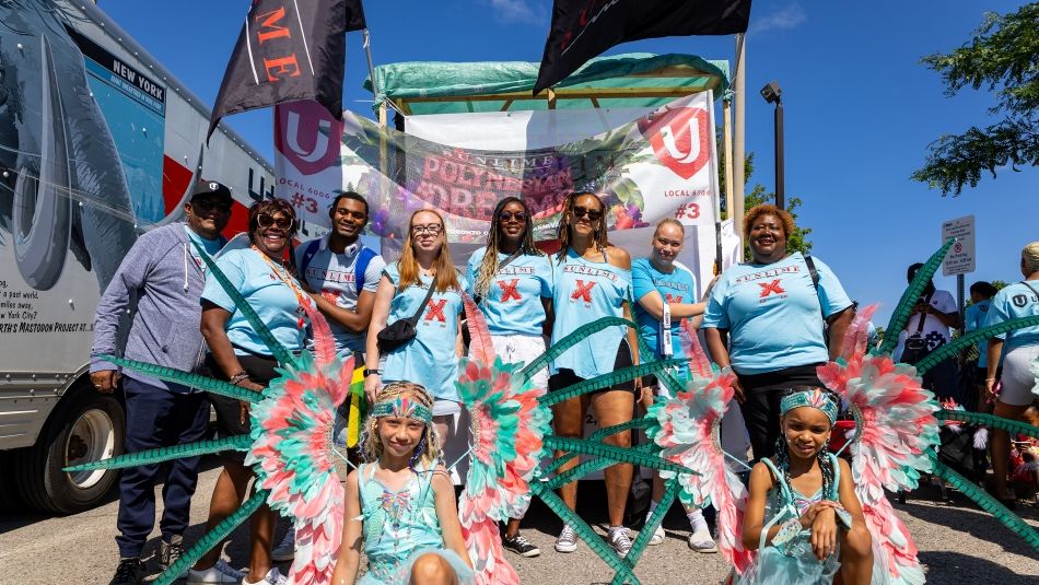 A group standing in front of a float and two young girls in front kneel wearing bright coloured carnival costumes 