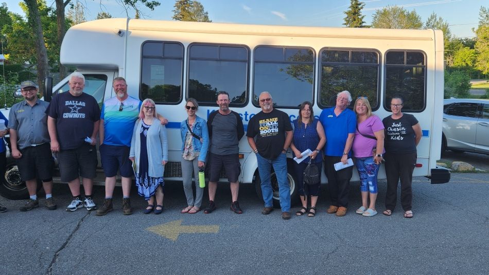 A group of 14 people standing in front of a parked white bus in a parking lot, with trees and other vehicles visible in the background.