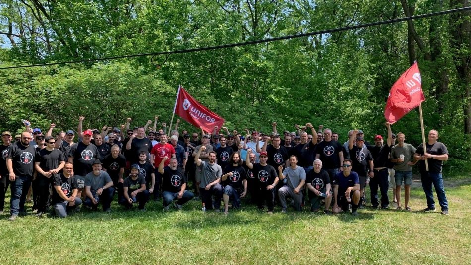 A group of people with their fists in the air, some holding red Unifor flags, kneeling on the ground and with trees in the background.