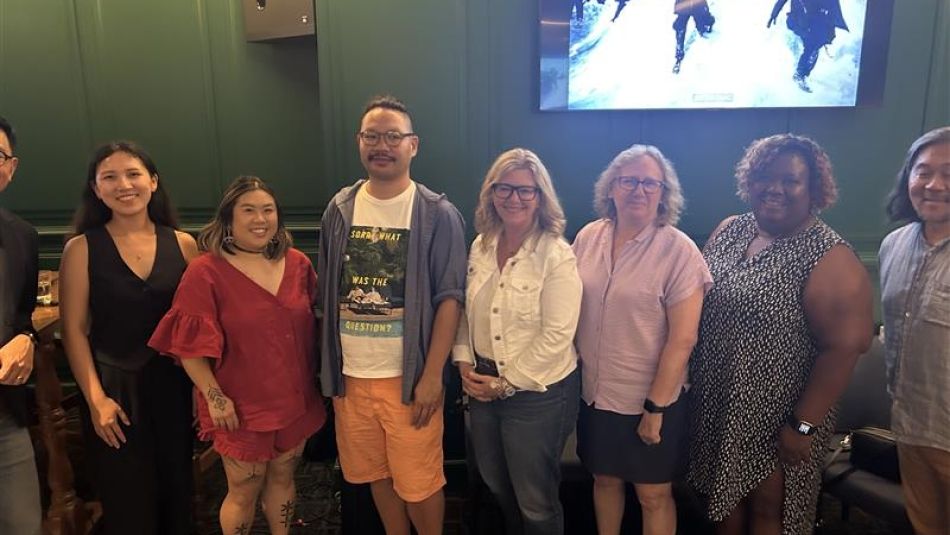 Seven people pose in front of a green wall with a screen hanging in the background of people climing on snow.