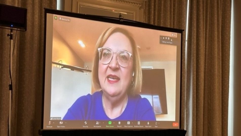 A woman with a blue shirt and glasses on a Zoom call projected on to a screen.