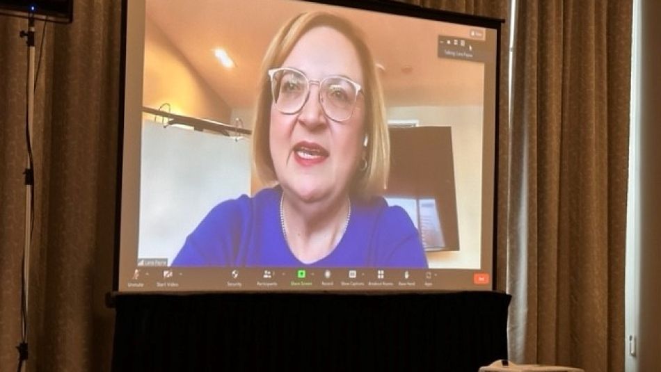 A woman with a blue shirt and glasses on a Zoom call projected on to a screen.