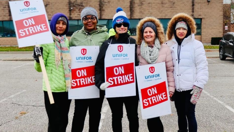Five women standing together outside holding red and white placards that read on strike with the Unifor logo displayed on them in front of a the Magna building in Windsor, Ontario.