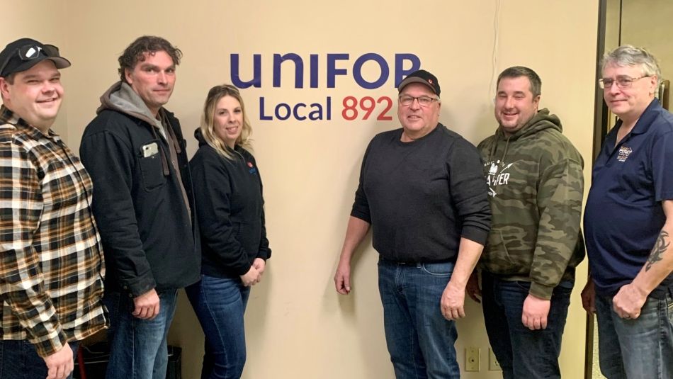 Eight Local 892 bargaining committee members posing in front of a Unifor logo decal on a wall