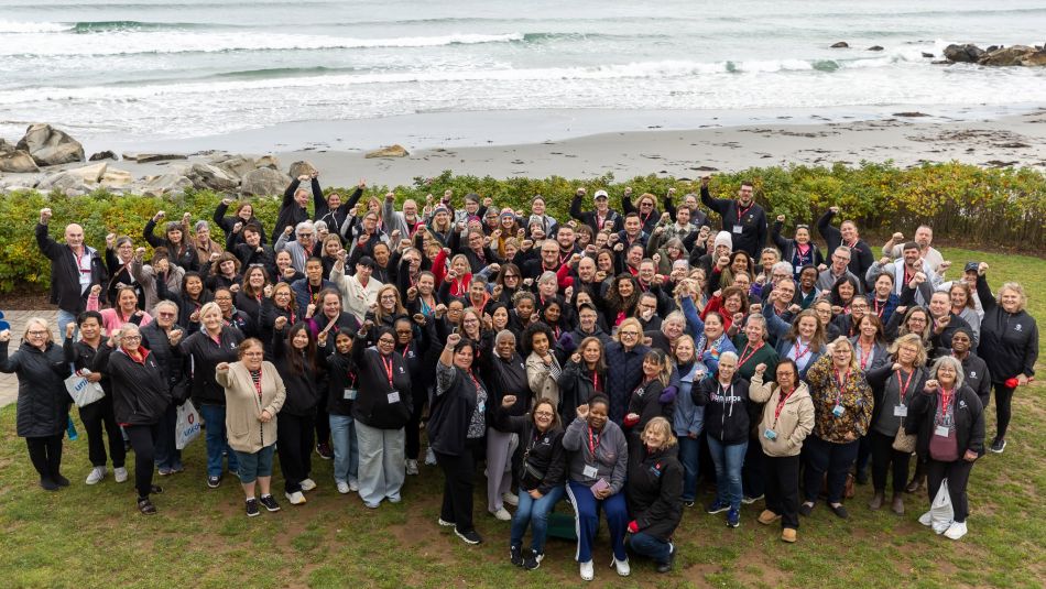 Large group of Unifor members posing outdoors beside a rocky beach, many raising their fists, with ocean waves in the background.