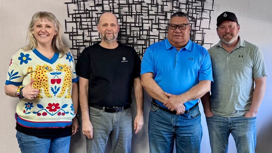 Quatre personnes se tiennent près d'un mur à l'intérieur. L'une d'elles fait le signe du « pouce levé ».