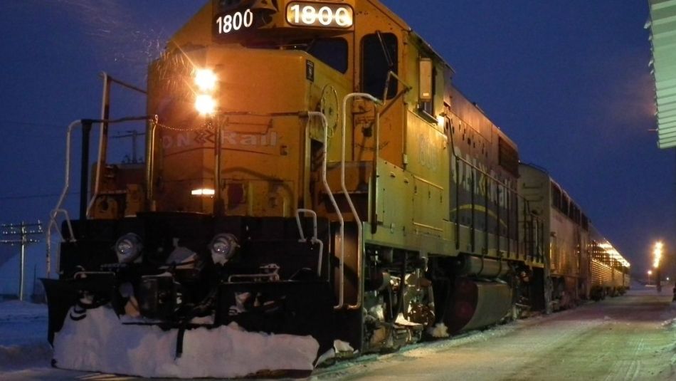 Ontario Northlander parked at a train station during twilight in a snowy setting. The locomotive's headlights and station lighting cast a warm glow on the surrounding area, contrasting with the blue hues of the early evening sky.