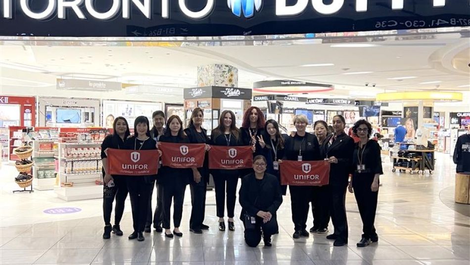 Thirteen women dressed in all black hold up red Unifor placards in front of a Toronto Duty Free store