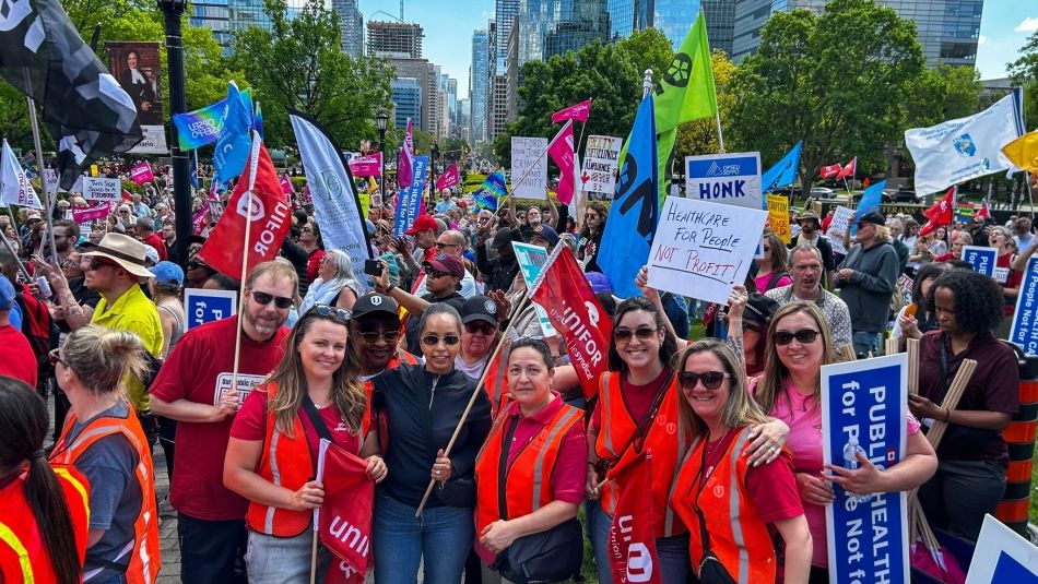 Unifor members pose for photo in front of large crowd holding signs about protecting public health care.