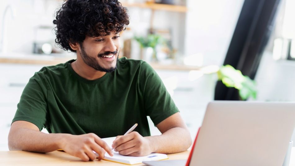Smiling man writing on notepad while looking at laptop