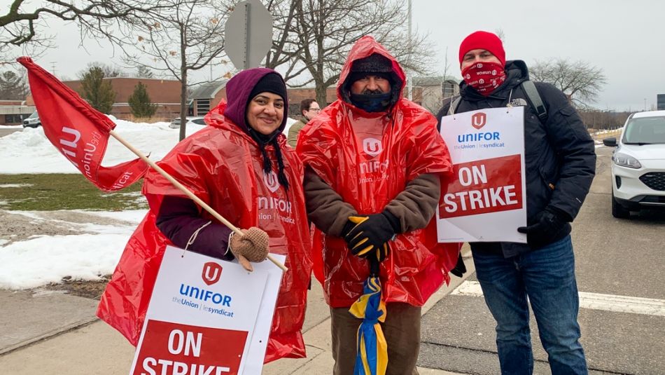 Three workers posing for a photo outside with a Unifor flag and On Strike signs