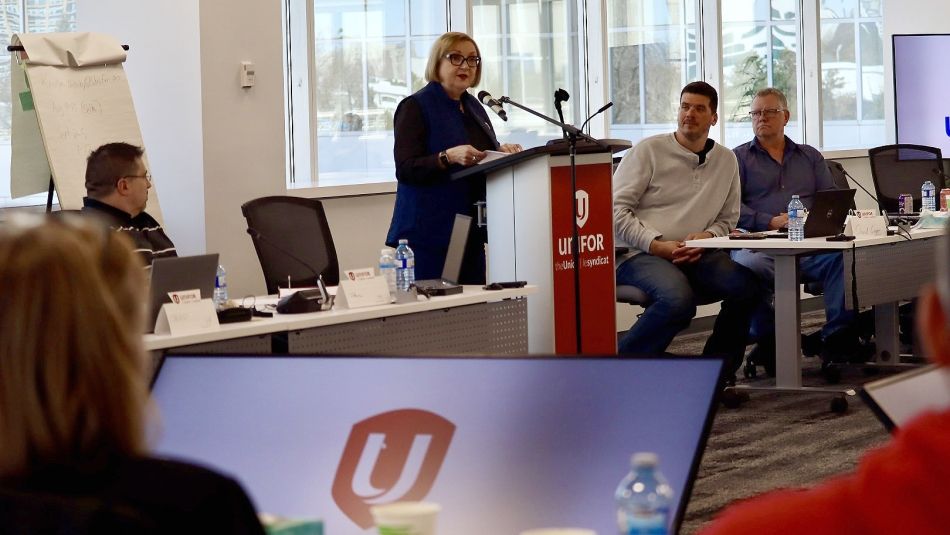 A women speaking at a podium with people sitting at tables to the side. A screen shows a Unifor shield.