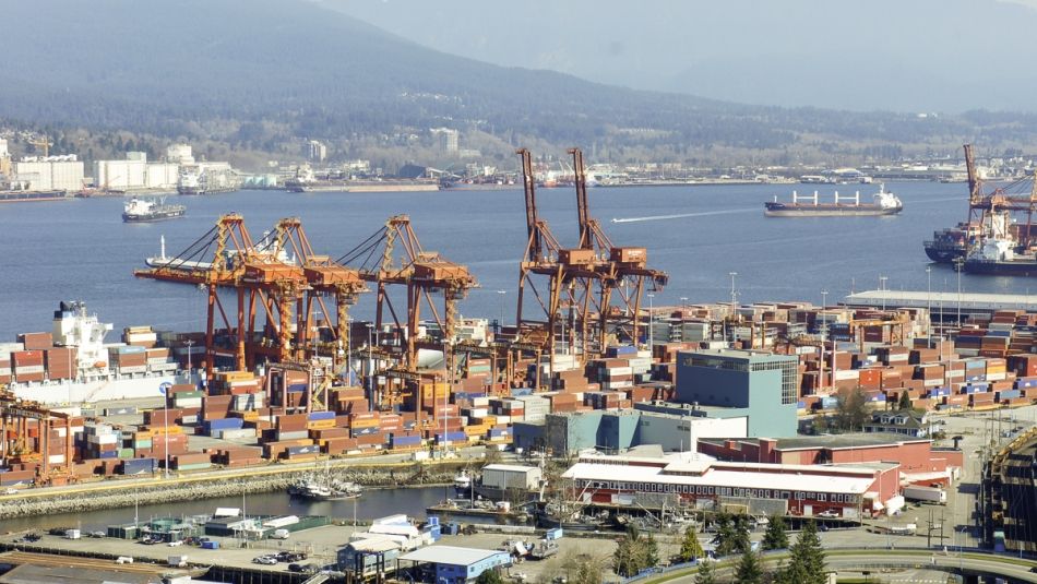 arial shot of a port with several large orange cargo cranes and a river and adjacent shore in the distance.