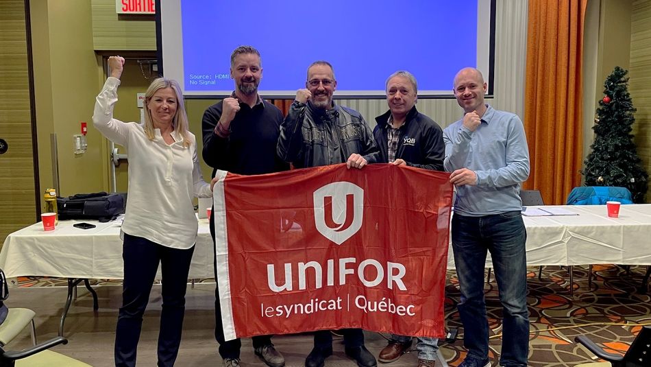 One woman with her arm raised in the air, stands with four men, all holding a red Unifor flag, with a table and projection screen in the background.