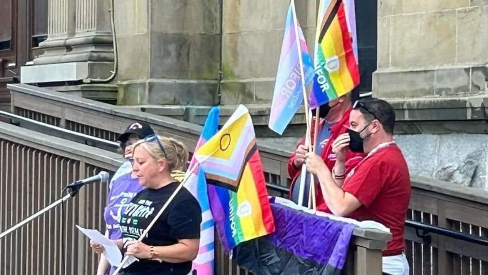 A wonen speaking at a mic with three people holding trans flags standing behind her.