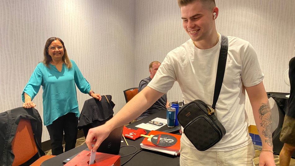 member putting a ballot into a ballot box with a woman watching from behind the table