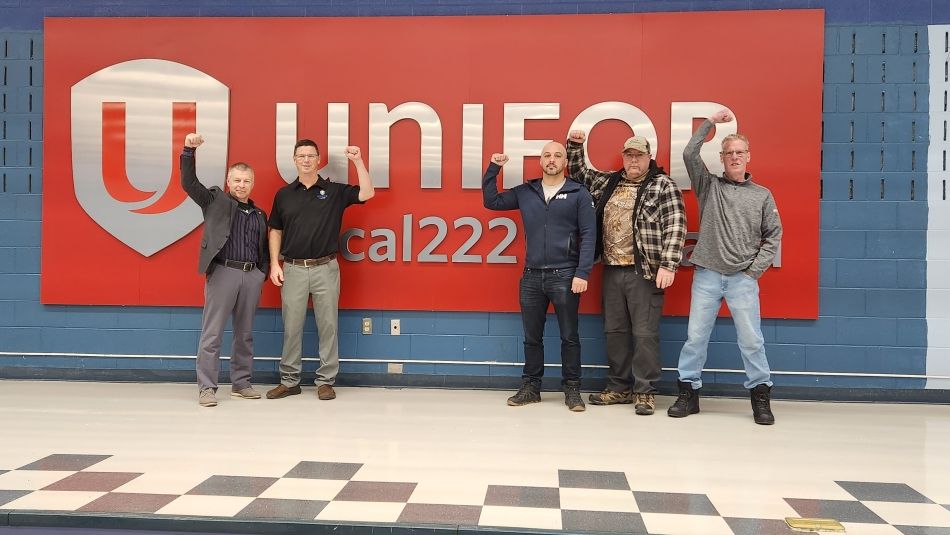 Five men with their fists raised in the air, standing in front of a large Unifor Local 222 red sign with a blue wall in behind them.