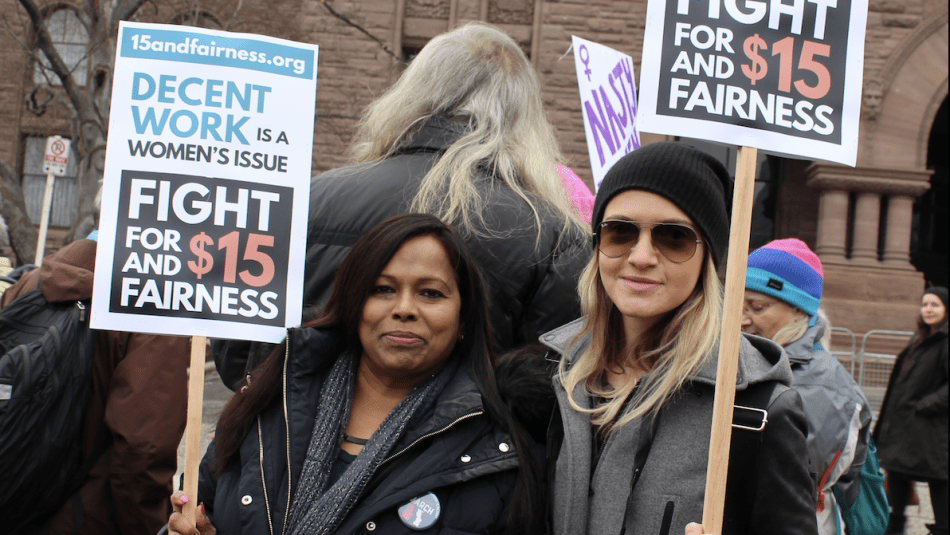 two women in front of the Legislative Building holding $15 for fairness signs