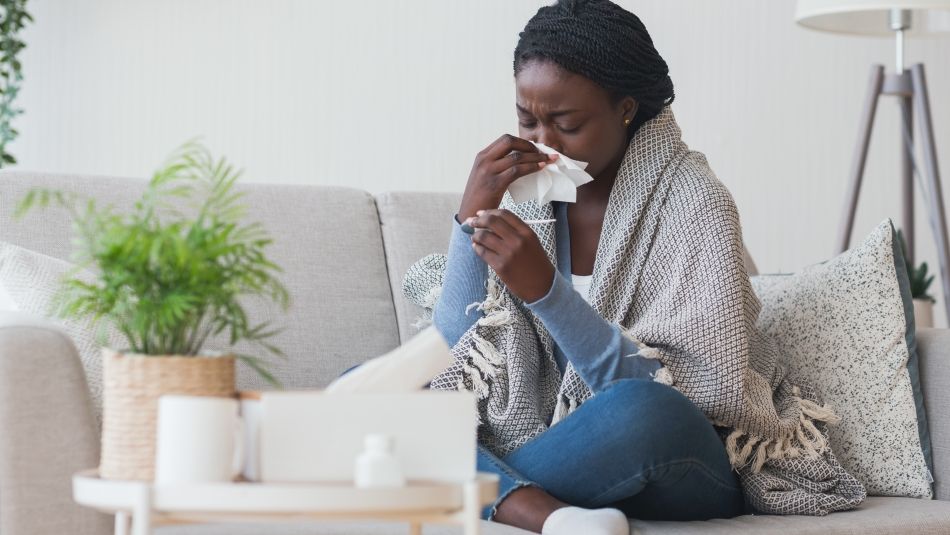 Woman on couch wiping nose with tissue and looking at thermometer.
