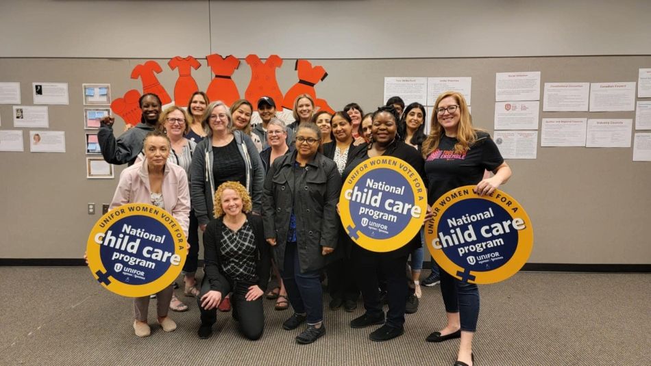 A group of women holding circular National Child Care Program signs. In the background are drawn oranges dresses cut out and papers stapled to the walls.