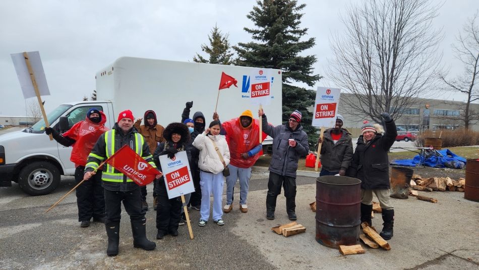  A group of people holding Unifor On Strike signs and flags at a picket line with white truck in the background.