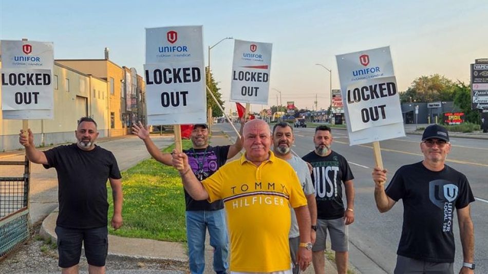 Six men stand on a sidewalk holding signs that read "UNIFOR LOCKED OUT," indicating a labor dispute.