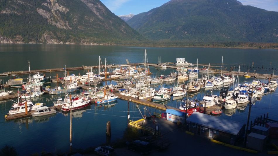 “A marina with fishing vessels on a sunny day. Mountains and water in the background.”