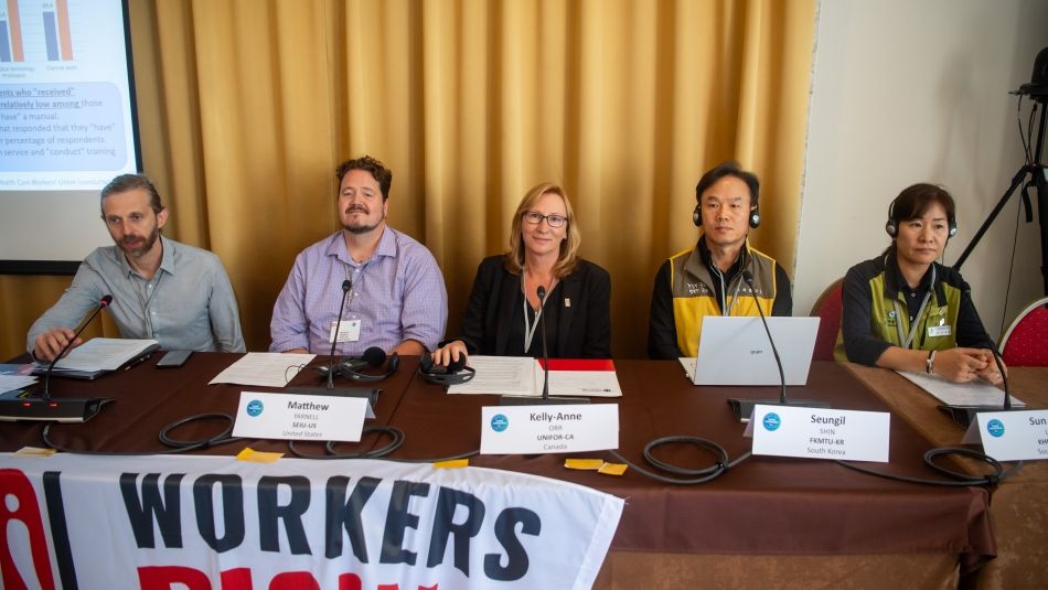 Five individuals are seated behind microphones at a table with a “Workers Rising” banner in front.