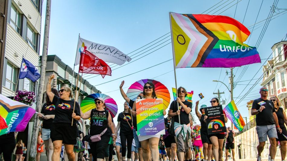 A group of people march down a city street holding pride flags and signs supporting unions and LGBTQ+ rights.