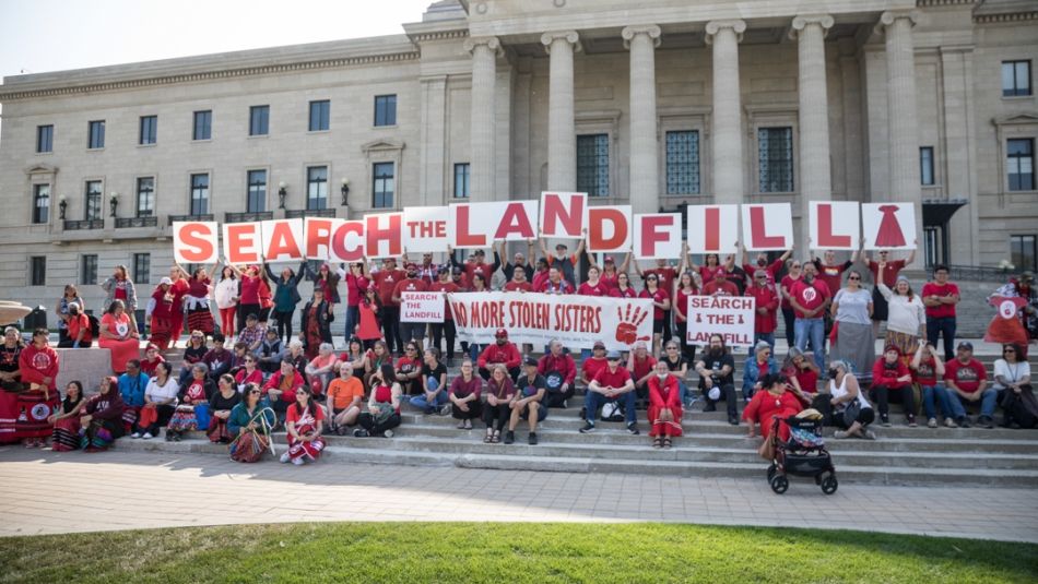 A large crowd wearing red and holding up signs that say Search the Landfill on the steps of the Manitoba Legislature. 