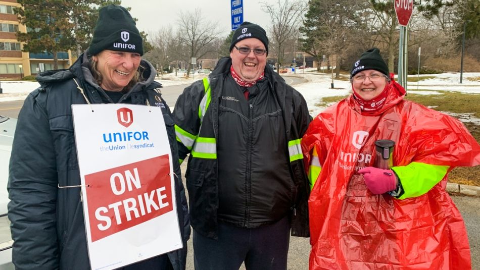 •	Trois travailleurs des transports en commun, tout sourire, tiennent une affiche « En grève » sur la ligne de piquetage