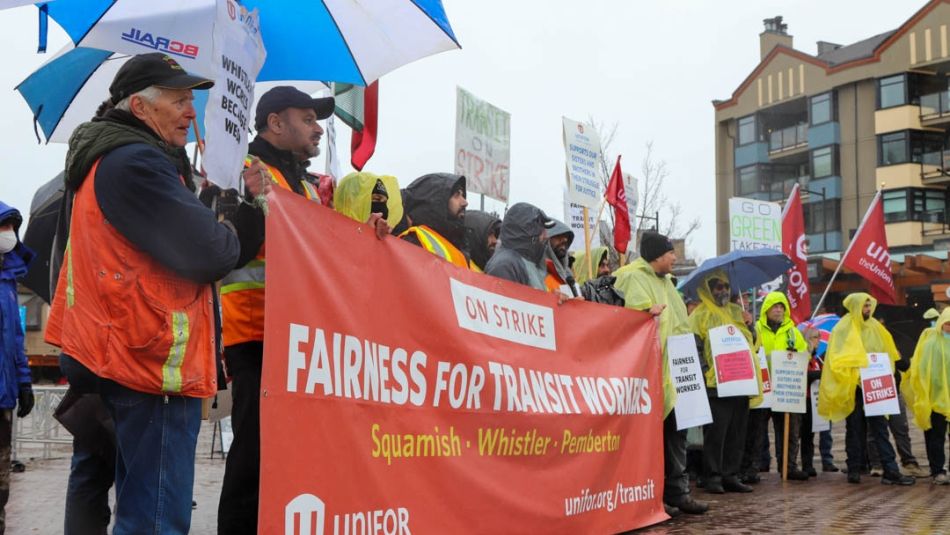 Une rangée de travailleurs du transport en commun à l’extérieur sous la pluie tenant une grande banderole rouge avec le slogan “Équité pour les travailleurs du transport en commun”