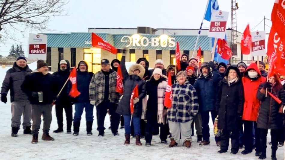 Grand groupe de travailleurs à l'extérieur avec des pancartes "En Greve" devant un bâtiment de Ro-Bus.