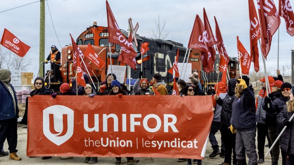 Unifor members holding flags stand behind a large red Unifor banner with a stopped CN train in background.