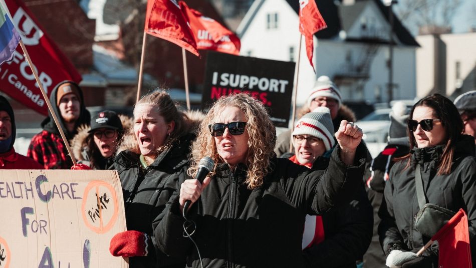 A woman wearing a black coat, shouting, with her arm in the air with people behind her, waving red Unifor flags.