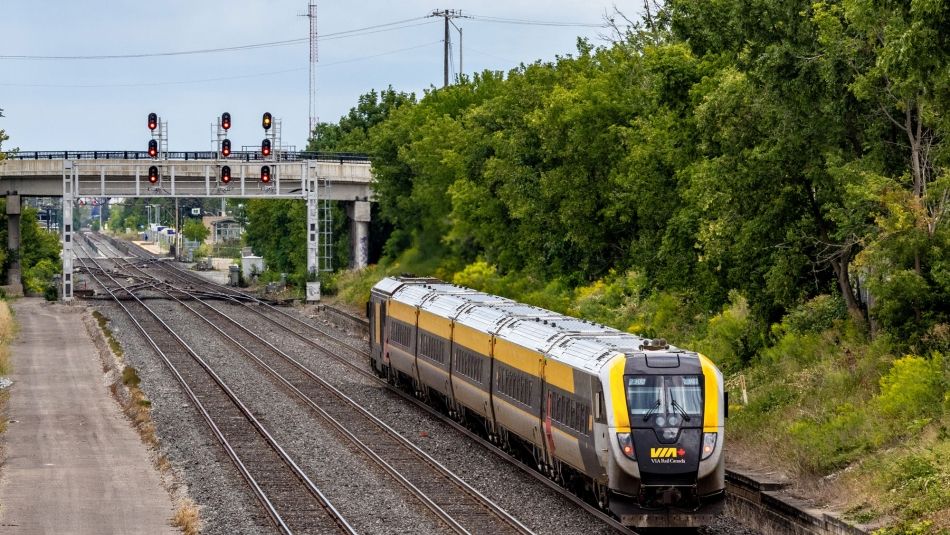 A Via Rail sleek modern Siemens train set with a signal gantry and road bridge in the distance.