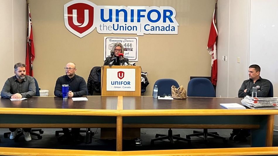 A woman speaks at a podium while three men are seated on either side. A Unifor sign is overhead.