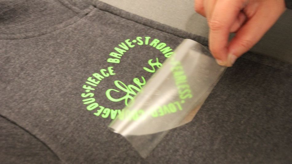 A hand carefully peals protective plastic away from a logo printed on a grey hoodie.