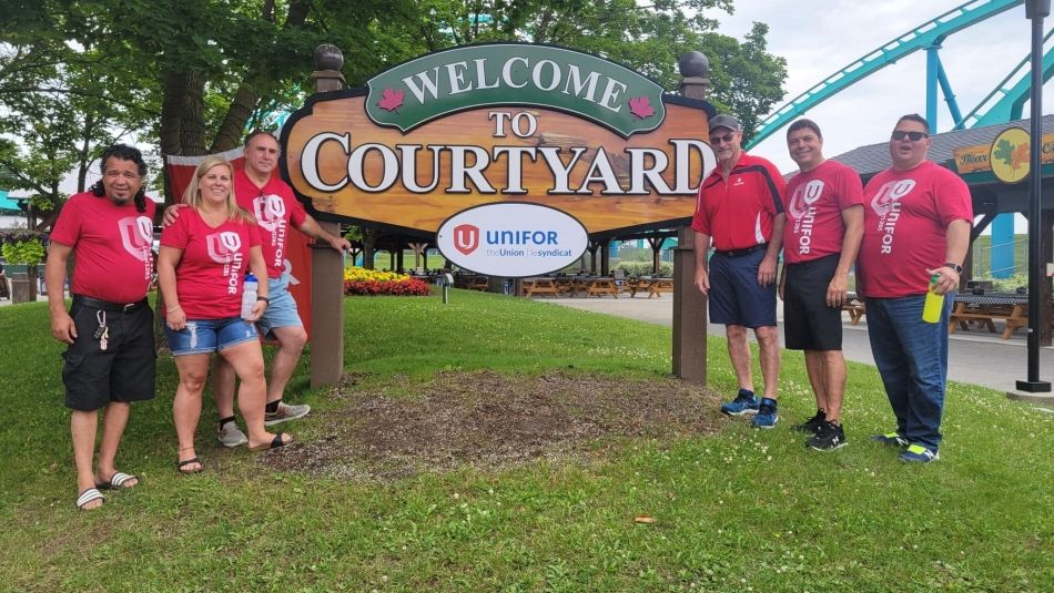 Six people wearing red Unifor shirts stand beside a sign that says “Welcome to Courtyard” with a Unifor sign below.