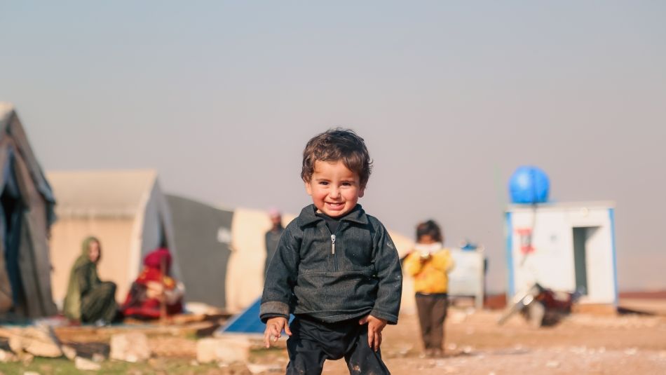 A young child smiles while walking through a refugee camp, with temporary shelters and families visible in the background, highlighting the resilience and humanity of people displaced from their homes.